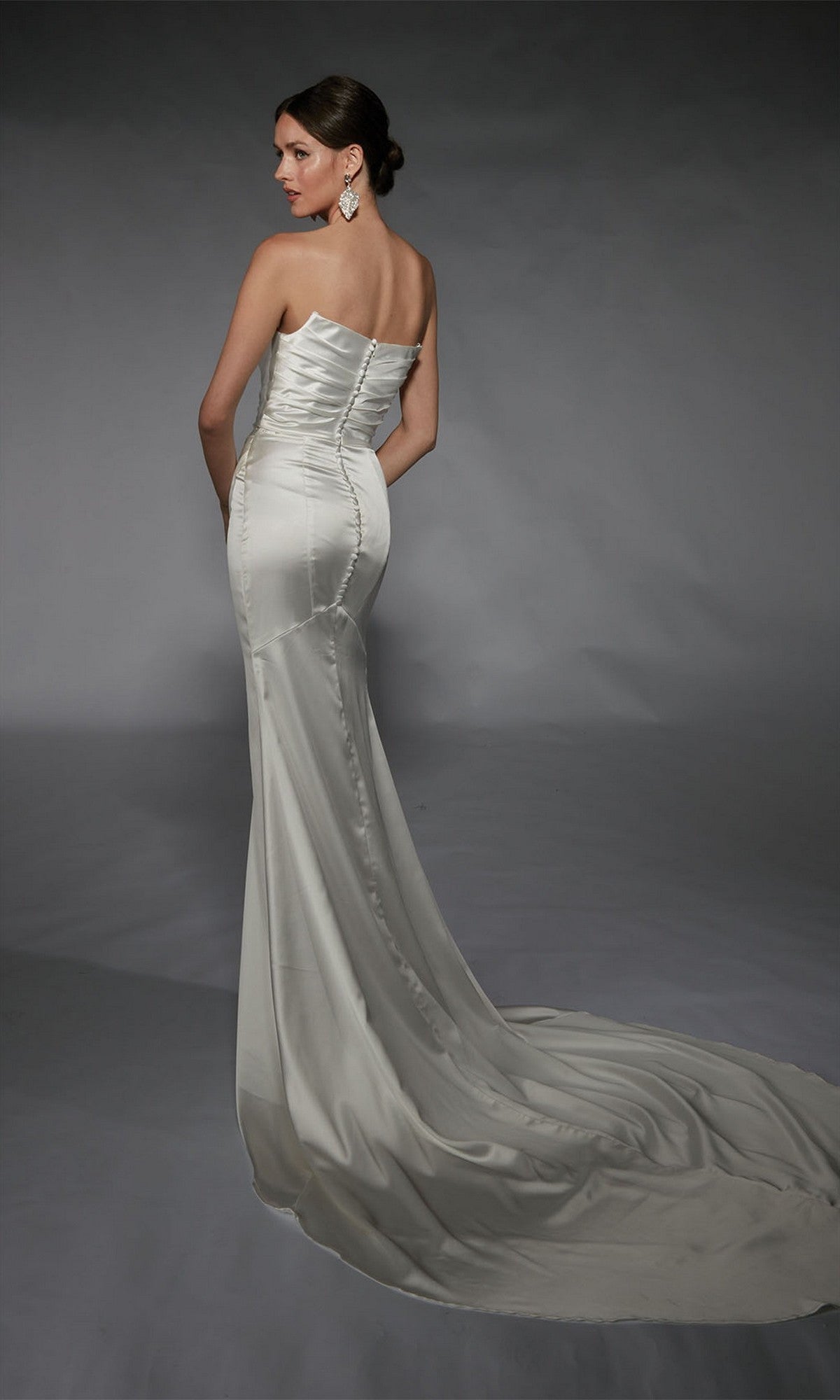 Woman in the Alyce 7106, a strapless fitted long white dress with a train, stands against a plain gray background, facing away from the camera.